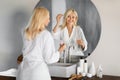 Elegant senior woman in bathrobe applying facial serum near mirror in bathroom Royalty Free Stock Photo