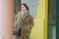 An elegant stylish girl in a beige fur coat, with a black bag, stands near a colored brick wall. A young girl with flowing hair Royalty Free Stock Photo