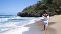 An elegant tourist in a white summer dress and a pink hat walks along the beach on a sunny day on the ocean. He looks