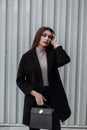 Elegant urban model young woman with long hair in shirt in luxurious fashionable black coat with leather handbag posing near metal