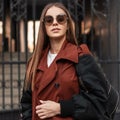 Elegant urban young hipster woman in stylish sunglasses in fashionable brown trench coat with trendy leather black backpack walks Royalty Free Stock Photo