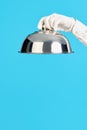 Elegant waiter& x27;s hand in white glove holding metal cloche lid cover on blue background Royalty Free Stock Photo