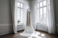 An elegant white wedding dress in a bright room with large windows, sheer curtains, wooden floor, and a floral touch