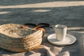 Elegant wicker straw bag on beige towel in sunlight. Cup of coffee, drink and plate. Concrete ground, sand beach