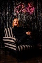 Elegant woman in black clothes sits in a chair. Party lettering on the wall. Red hair. Bright, shiny background