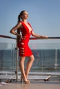 An elegant woman on a light sea background. A beautiful lady in a bright red dress with a cleavage. A girl posing on a terrace. Royalty Free Stock Photo