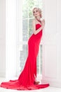 Elegant woman with perfect makeup and red long dress, posing near window. Royalty Free Stock Photo