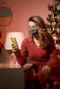 Elegant woman in a red cocktail dress, wearing protective face mask by the christmas tree, alone, lonely, talking and writing on Royalty Free Stock Photo