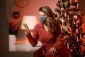 Elegant woman in a red cocktail dress, wearing protective face mask by the christmas tree, alone, lonely, talking and writing on Royalty Free Stock Photo