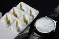 An elegant wristwatch next to medical yellow pill capsules in a silver blister and white box. Prescription and Schedule Medication