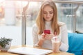 Elegant young blonde woman in white sweater sits on blue sofa in cafe, holds pink mobile phone and surfes internet or types mesage