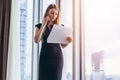 Elegant young female business owner holding documents talking on mobile phone discussing contract conditions standing in Royalty Free Stock Photo