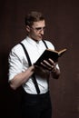 A man in round glasses, suspenders, a white shirt is reading a book