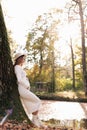 Elegant young pregnant woman in white knit dress and hat touching and stroking belly, relaxing in park by the lake on Royalty Free Stock Photo