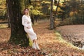 Elegant young pregnant woman in white knit dress and hat touching and stroking belly, relaxing in park by the lake on Royalty Free Stock Photo