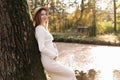 Elegant young pregnant woman in white knit dress and hat touching and stroking belly, relaxing in park by the lake on Royalty Free Stock Photo