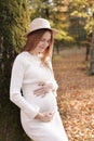 Elegant young pregnant woman in white knit dress and hat touching and stroking belly, relaxing in park by the lake on Royalty Free Stock Photo