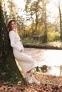 Elegant young pregnant woman in white knit dress and hat touching and stroking belly, relaxing in park by the lake on Royalty Free Stock Photo