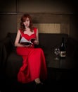 Elegant young redhead woman in a red dress, reads glamor magazine, having a glass of red wine Royalty Free Stock Photo