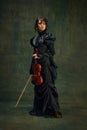 Elegant young woman in black dress, professional violinist standing with violin against vintage green background