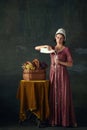 Elegant young woman in renaissance dress, maid pouring milk into glass against dark vintage background. Peasant girl Royalty Free Stock Photo