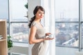 Elegant young woman standing in a spacious light studio, drinking cup of coffee, smiling, dreaming near large window Royalty Free Stock Photo