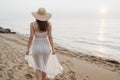 Elegant young woman with white dress and sunhat walking by the beach in a warm sunset Royalty Free Stock Photo