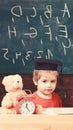 Elementary education concept. Pupil in mortarboard, chalkboard on background. Kid studies near alarm clock and teddy