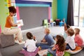 Elementary Pupils Raising Hands To Answer Question As Female Teacher Reads Story In Classroom Royalty Free Stock Photo