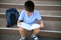 Elementary student doing homework sitting next to his school bag on the stairs of school establishment. Back to school