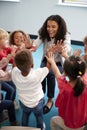 Elevated view of infant school children in a circle in the classroom giving high fives to their smiling female teacher, vertical,