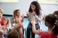 Elevated view of infant school children sitting on chairs in a circle in the classroom, raising hands and learning to count with t