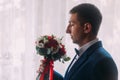 Ellegant bridegroom with bow-tie holding a bouquet. White curtains are in the background