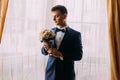 Ellegant dressed romantic groom holding a wedding bouquet of pale pink roses in hands standing near the window