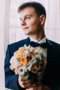 Ellegant dressed romantic groom holding a wedding bouquet of white and pale orange roses in hands standing near the