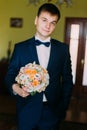 Ellegant dressed stylish groom holding a wedding bouquet of white and pale orange roses in hands standing in hotel room