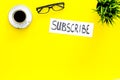 Email subscribe concept. Hand lettering subcribe on work desk with plant, glasses, cup of coffee on yellow background