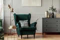 Emerald green armchair with pillow next to grey wooden commode in dark living room interior