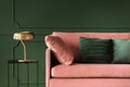 Emerald green pillows on pastel pink couch in stylish living room interior with dark green wall