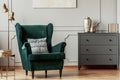 Emerald green wing back chair in grey living room interior with wooden commode Royalty Free Stock Photo