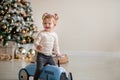 The emotion of a child. A little girl in a white sweater sits on a toy antique blue car and laughs. Christmas tree in the Royalty Free Stock Photo