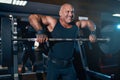 Emotional adult bald bodybuilder doing trapezius and deltoid exercise with a barbell in the gym Royalty Free Stock Photo