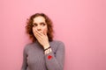 Emotional curly girl with shocked covered face looks away to empty space. Portrait of surprised red-haired girl stands on pink