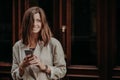 Emotional delighted happy brunette young woman with dark hair, uses mobile phone for texting messages, dressed in elegant raincoat
