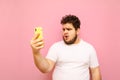 Emotional fat young man in white t-shirt is using smartphone on pink background, with shocked face looking into camera. Funny full Royalty Free Stock Photo