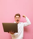 Emotional guy rejoices in victory with a laptop in his hands, looks into the camera and shouts with joy,isolated on pink