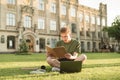 Emotional guy sitting with books and a laptop on a lawn on the background of a university building,reading a notebook with