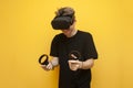 Emotional guy in VR glasses plays a shooter on a yellow background, a gamer shoots with virtual weapons and holds modern joysticks