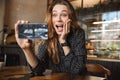 Emotional happy beautiful young pretty woman sitting in cafe indoors using mobile phone talking with friends take a selfie Royalty Free Stock Photo