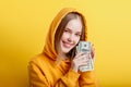 Emotional happy teenage blonde girl in hood win money cash holding dollars in hands isolated on color yellow background. Portrait Royalty Free Stock Photo